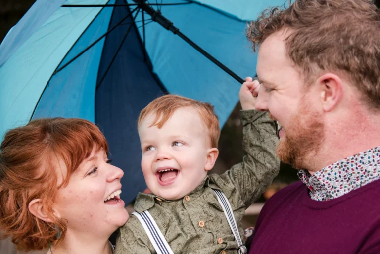 two parents and their little boy playfully look at each other under a blue umbrella - shot at Gage park in Hamilton, Ontario