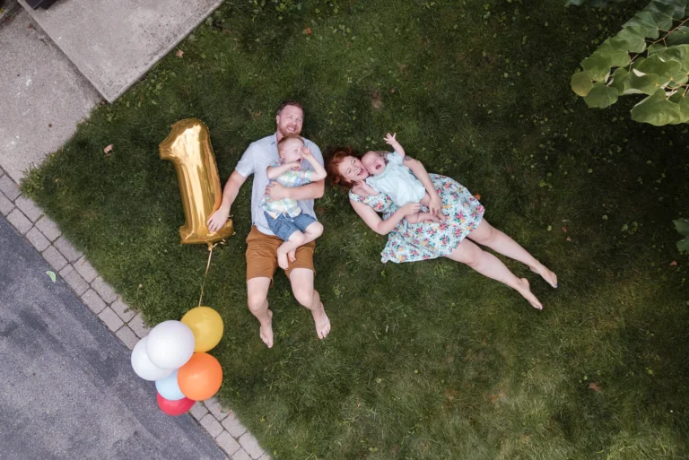 a drone shot looking down at a family of four laying in their front yard while dad holds a 1 mylar balloon for the baby's first birthday