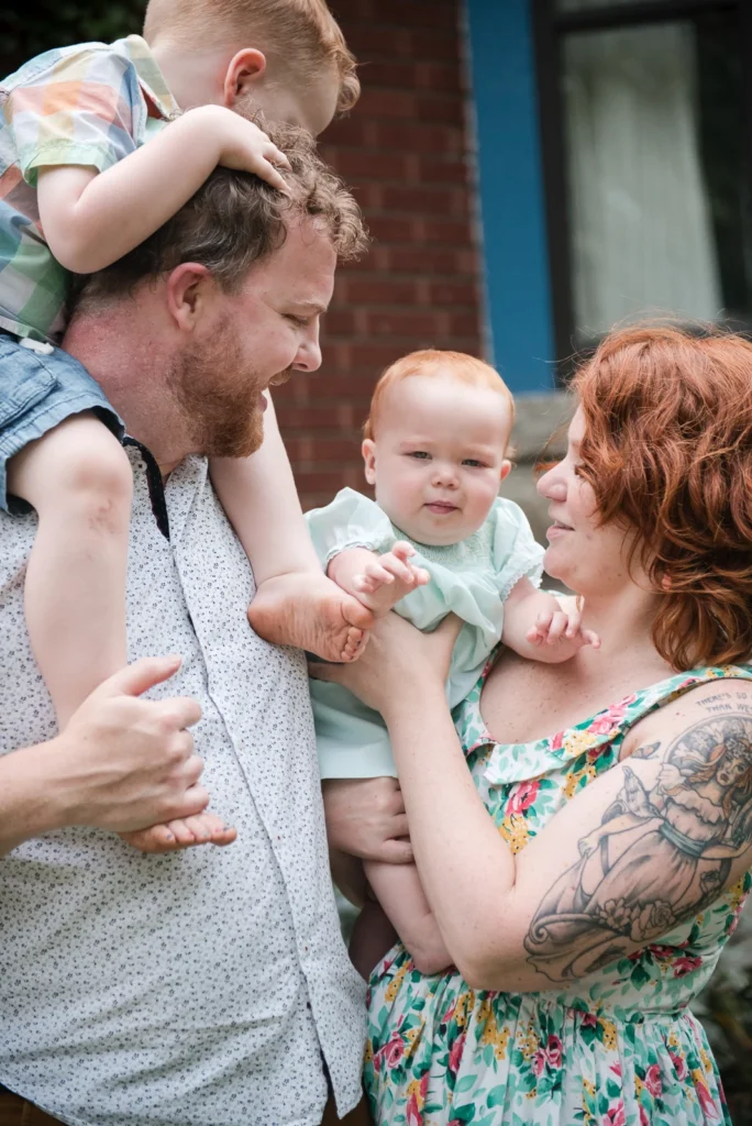 a family of four, a toddler boy on dad's shoulders while mom holds the baby who is looking at the camera