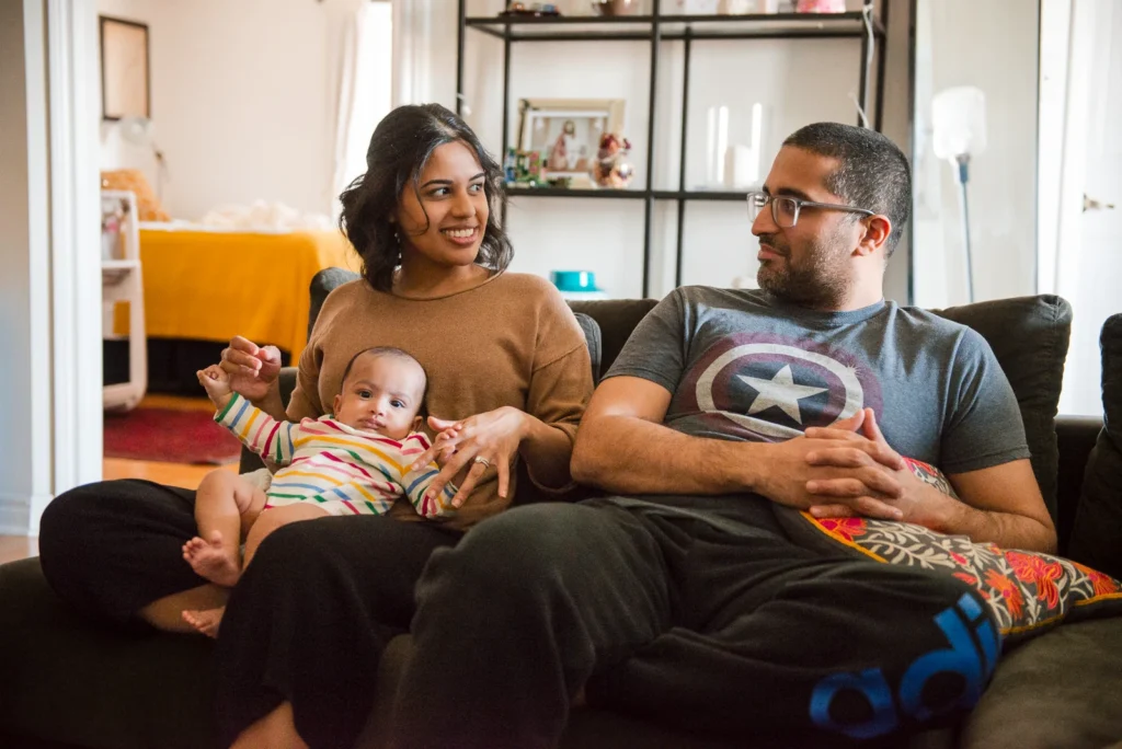 a couple look at each other while sitting on the sofa holding their baby - an in home family portrait shot in Hamilton, Ontario