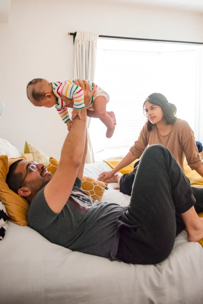 a father holds his baby above his head in bed as the mother looks on - an in home family portrait shot in Hamilton, Ontario