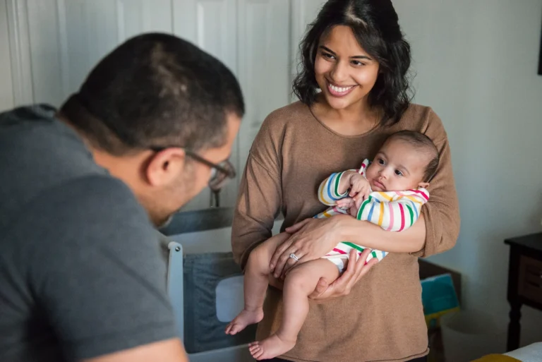 a woman holds her baby as the father playfully looks at the child - an in home family portrait shot in Hamilton, Ontario