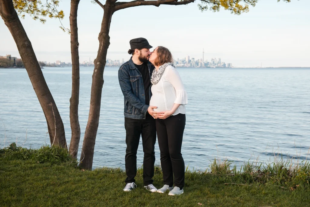 a pregnant woman and her partner shares a kiss with the toronto skyline in the background - a family portrait shot in Hamilton, Ontario