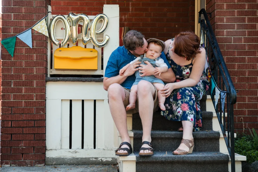 a couple kisses their baby boy as they sit on their porch stairs with a first birthday balloon nearby - a family portrait shot in Hamilton, Ontario