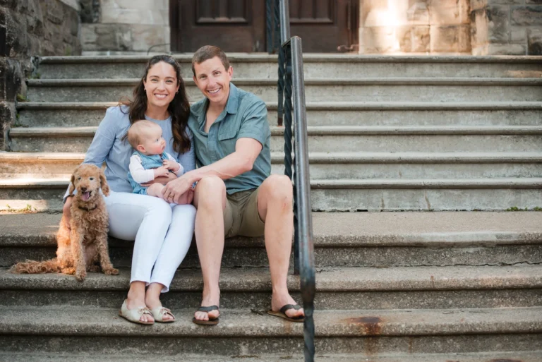 a couple sit on stone stairs and smile at the camera as they hold their baby and dog - a family portrait shot in Hamilton, Ontario