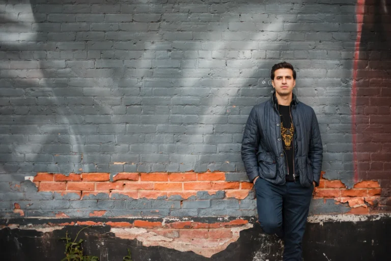 a man stands against a rustic wall and looks at the camera - a headshot shot by Hamilton headshot photographer : Pixelesque Photography