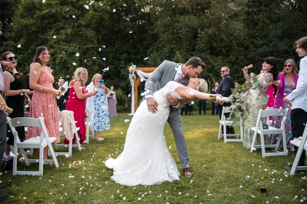 A couple share a dip and kiss at the end of the aisle as confetti flies around them. Shot by Pixelesque Photography - Hamilton Wedding Photographer.