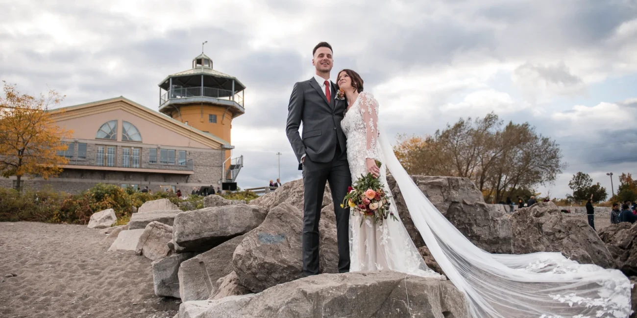 The wedding couple poses on rocks athe beach in front of a lighthouse. Shot at Lakeview by Carmens in Hamilton by Pixelesque Photography - Hamilton Wedding Photographer.