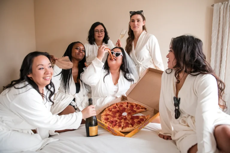 The bride and her bridesmaids enjoy pizza and champagne in their robes on a bed. Shot by Pixelesque Photography - Hamilton Wedding Photographer.