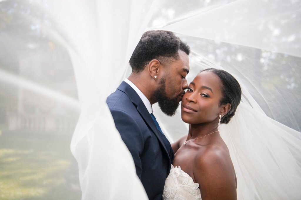 a groom kisses a bride's cheek under their veil as she looks at the camera. photographed at the guild inn estate by Pixelesque Photography - Hamilton Wedding Photographer