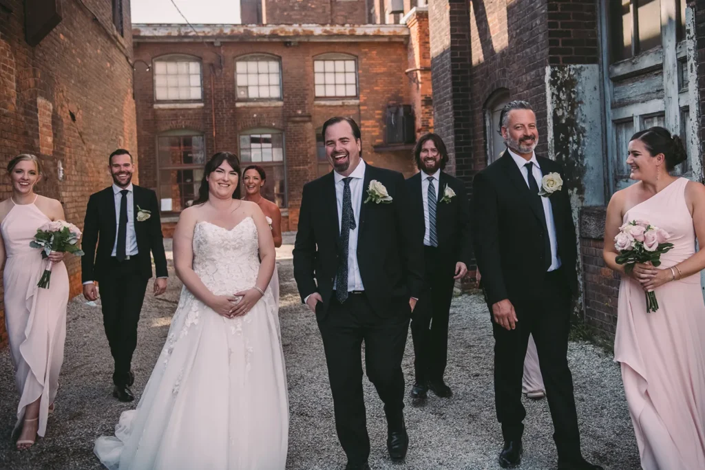 a bride, groom, and their wedding aprty laugh and walk along a rustic alley at the cotton factory - hamilton wedding venue - shot by hamilton wedding photographer- Pixelesque Photography