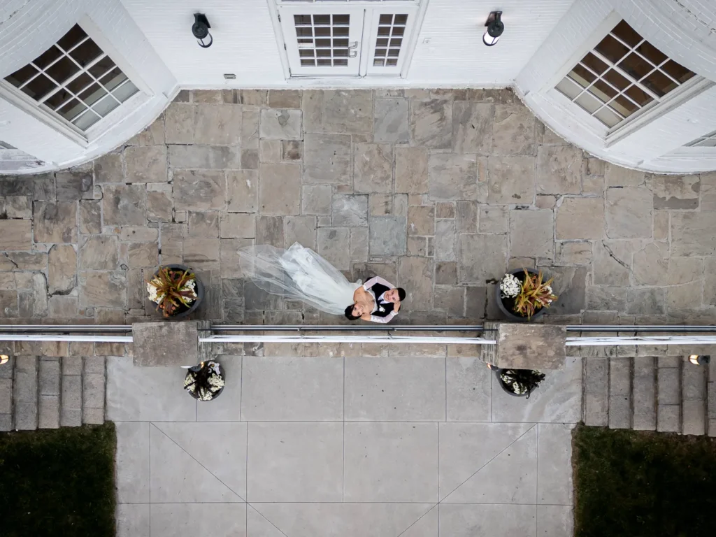 the couple looks up at the drone as they are in each others' arms. One of many beautiful moments from a wedding at Harding Waterfront Estate in Oakville, by Pixelesque Photography - Hamilton Wedding Photographer.