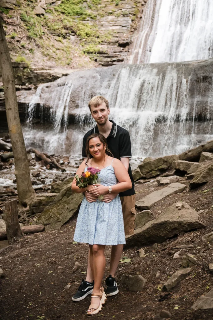 a couple share a hug in front of a waterfall. A couple stand and look at a raging waterfall. Engagement Session Shot at Sherman Falls, Ancaster, by Pixelesque Photography - Hamilton Wedding Photographer.