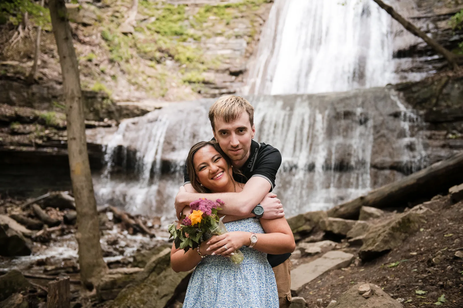 a couple embrace and laugh with a waterfall behind them. A couple stand and look at a raging waterfall. Engagement Session Shot at Sherman Falls, Ancaster, by Pixelesque Photography - Hamilton Wedding Photographer.