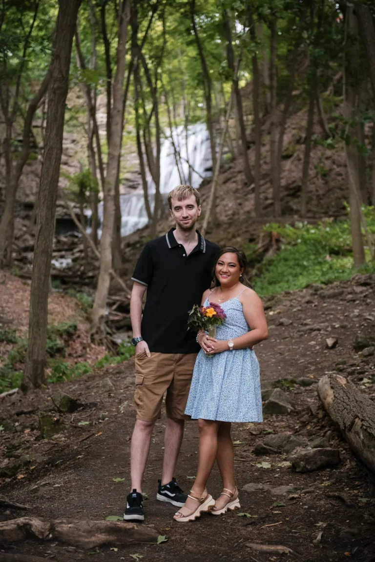 a couple stand beside each other in the woods. A couple stand and look at a raging waterfall. Engagement Session Shot at Sherman Falls, Ancaster, by Pixelesque Photography - Hamilton Wedding Photographer.