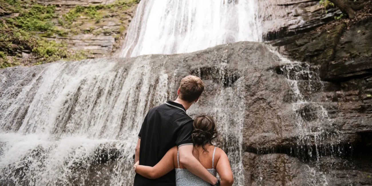 A couple stand and look at a raging waterfall. Engagement Session Shot at Sherman Falls, Ancaster, by Pixelesque Photography - Hamilton Wedding Photographer.
