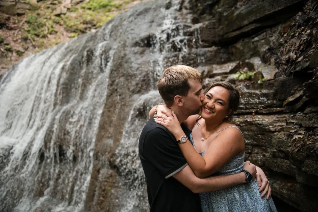a couple cuddle on a rock wall beside a waterfall. Engagement Session Shot at Sherman Falls, Ancaster, by Pixelesque Photography - Hamilton Wedding Photographer.