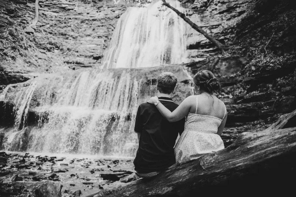 a rear view of a couple sitting on a log looking at a waterfall. Engagement Session Shot at Sherman Falls, Ancaster, by Pixelesque Photography - Hamilton Wedding Photographer.