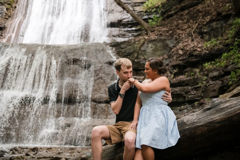 a couple sits on a log in front of a waterfall and the man holds and kisses her ring. Engagement Session Shot at Sherman Falls, Ancaster, by Pixelesque Photography - Hamilton Wedding Photographer.