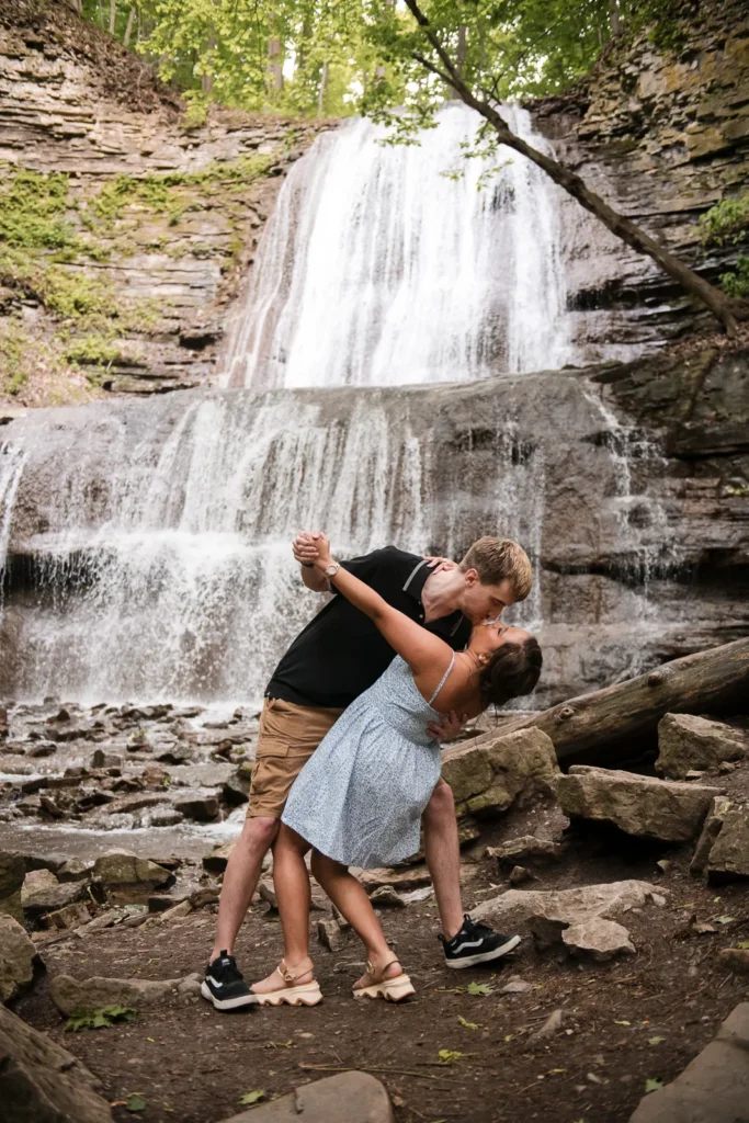a man dips and kisses his partner in front of a waterfall. Engagement Session Shot at Sherman Falls, Ancaster, by Pixelesque Photography - Hamilton Wedding Photographer.