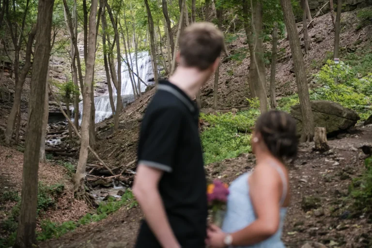 a couple look to the waterfalls in the distance. the couple is out of focus and the waterfall is in focus. A couple stand and look at a raging waterfall. Engagement Session Shot at Sherman Falls, Ancaster, by Pixelesque Photography - Hamilton Wedding Photographer.