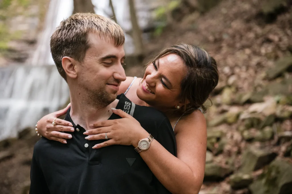 a woman hugs her partner from behind in the woods with a waterfall behind them. Engagement Session Shot at Sherman Falls, Ancaster, by Pixelesque Photography - Hamilton Wedding Photographer.