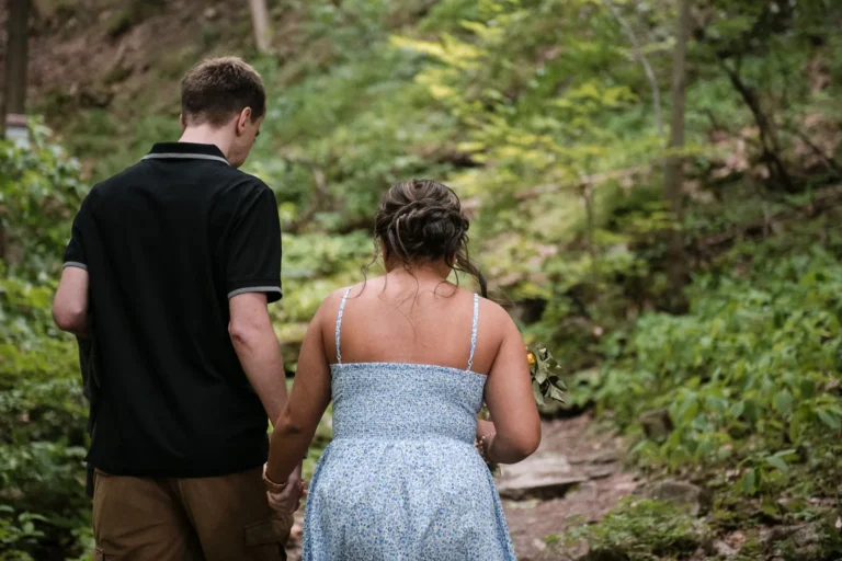 a couple walks hand in hand thorugh the woods. A couple stand and look at a raging waterfall. Engagement Session Shot at Sherman Falls, Ancaster, by Pixelesque Photography - Hamilton Wedding Photographer.
