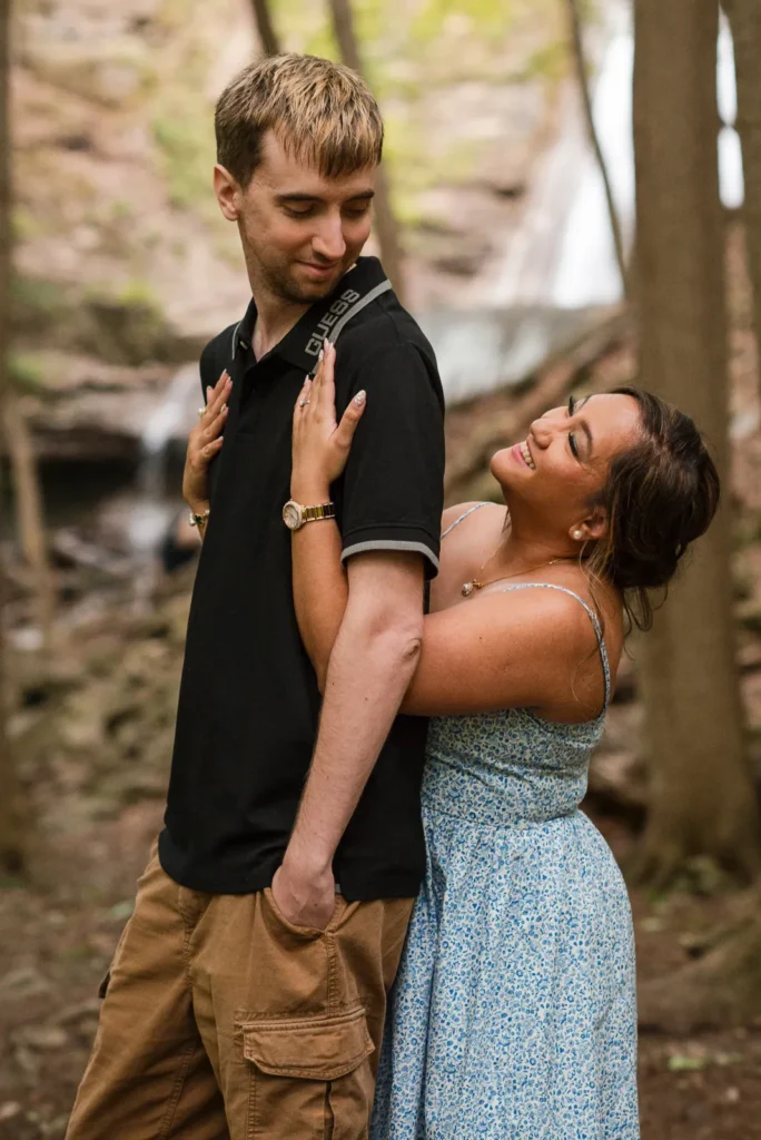 a woman hugs her partner from behind in the woods. Engagement Session Shot at Sherman Falls, Ancaster, by Pixelesque Photography - Hamilton Wedding Photographer.
