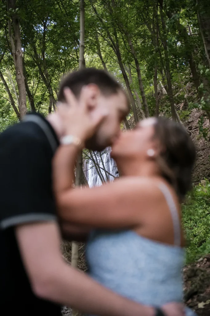 An out of focus couple kisses with a waterfall in focus behind them. Engagement Session Shot at Sherman Falls, Ancaster, by Pixelesque Photography - Hamilton Wedding Photographer.