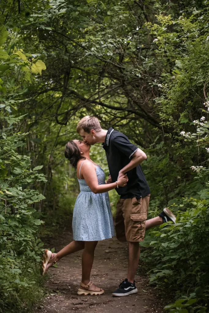 A couple shares a kiss in the woods as they both lift a leg behind them. Engagement Session Shot at Sherman Falls, Ancaster, by Pixelesque Photography - Hamilton Wedding Photographer.