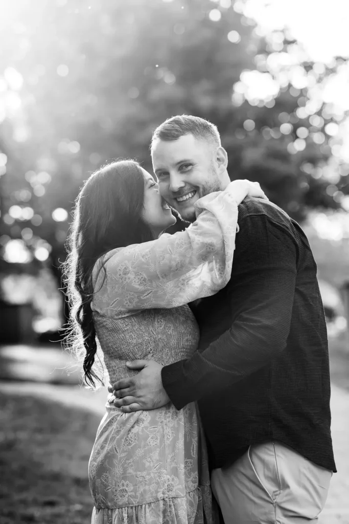 A black and white image of a couple embracing as the man looks at the camera and the woman kisses his cheek in Simcoe Park. Shot in Niagara-on-the-lake by Pixelesque Photography.