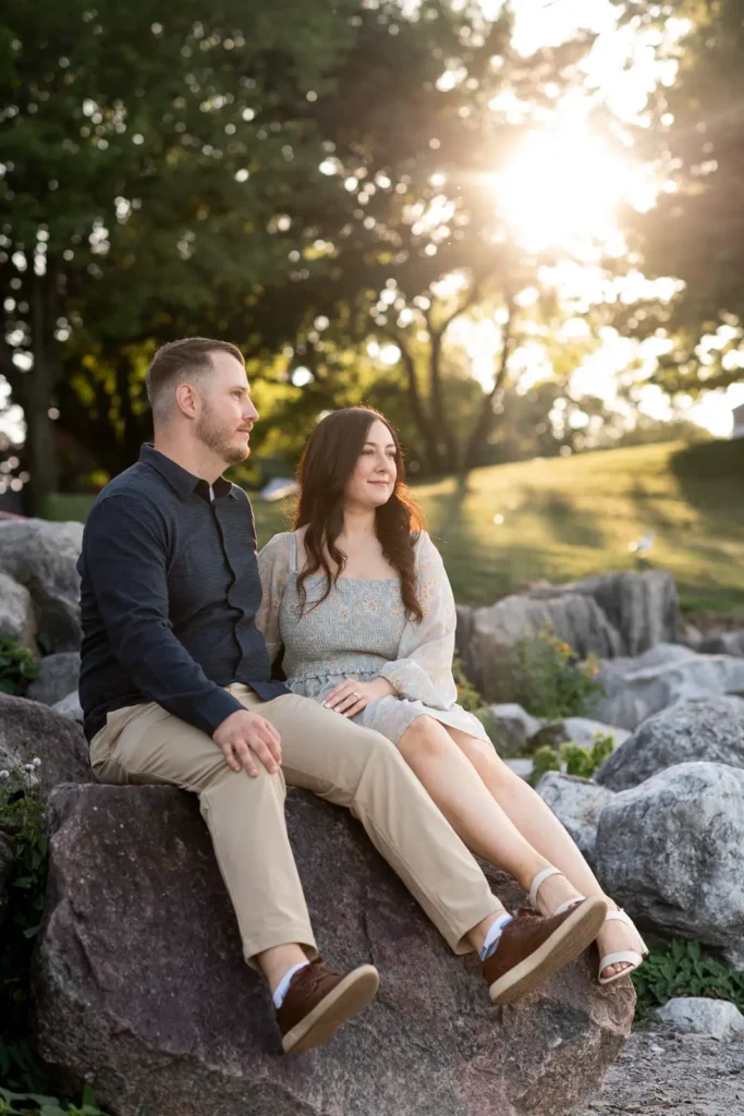 a couple sit beside each other on the rocks and look out to the water as the sun bursts behind them in Queen's Royal Park. Part of an engagement shoot Shot in Niagara-on-the-lake by Pixelesque Photography.