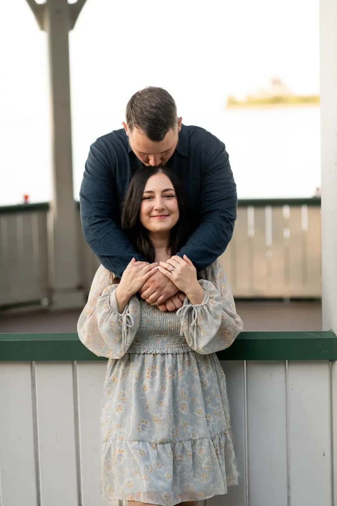 a man kisses the top of his partner's head as she stands outside a gazebo and he is inside. Part of an engagement shoot Shot in Niagara-on-the-lake by Pixelesque Photography.