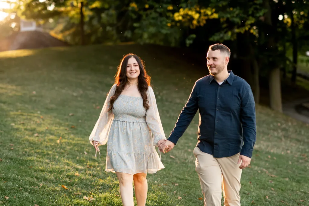 a couple walk in the grass as the sun shines on them in the Queen's Royal Park. Part of an engagement shoot Shot in Niagara-on-the-lake by Pixelesque Photography.