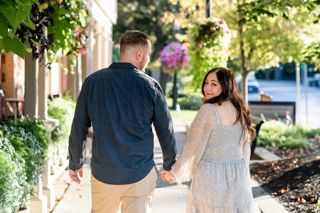 a couple holds hands and walks down a quaint street as the woman looks back at the camera. Shot in Niagara-on-the-lake by Pixelesque Photography.