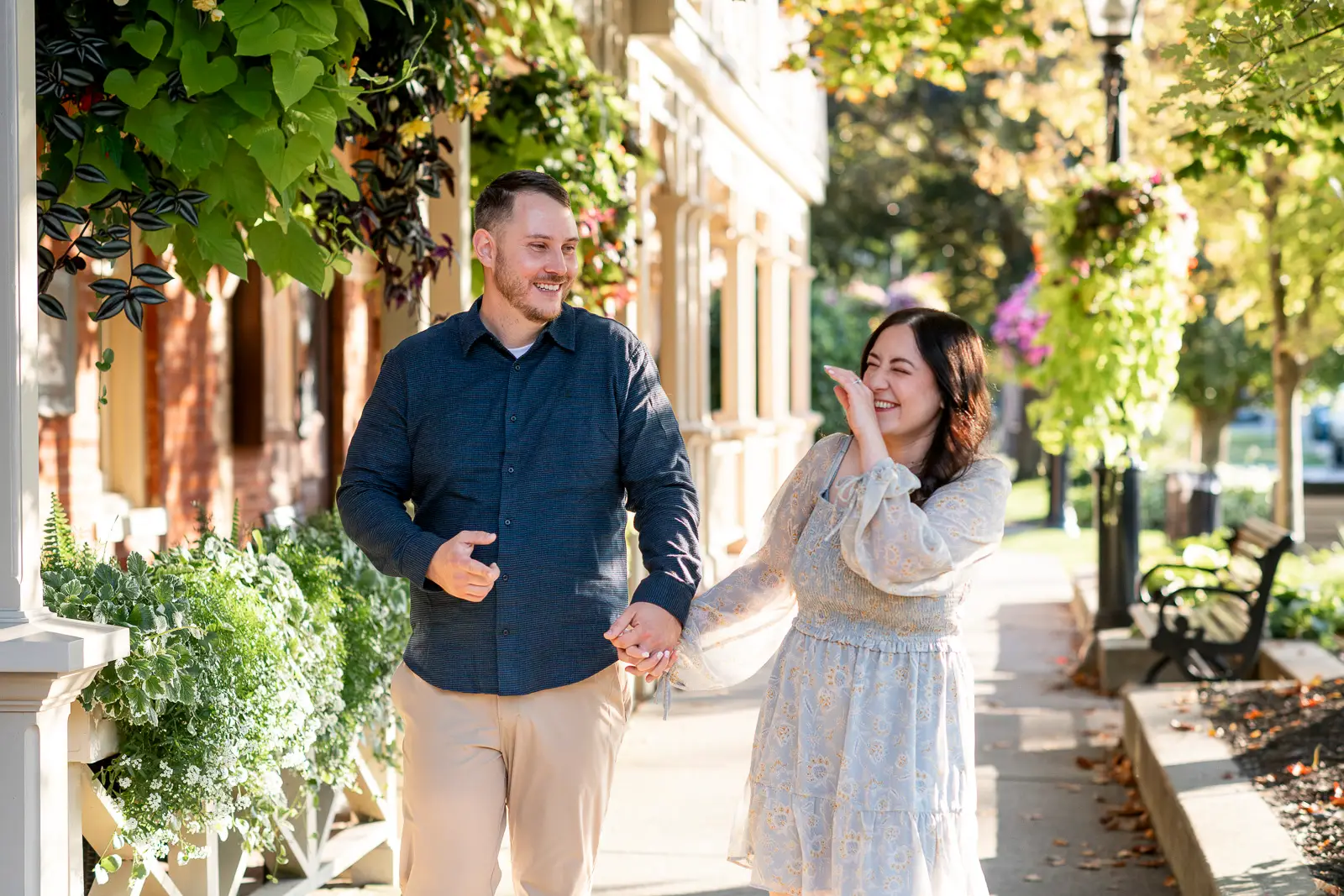 a couple laughs as they walk through a quaint street surrounded by foliage. Rachel & Matt’s Romantic Engagement Session in Niagara-on-the-Lake