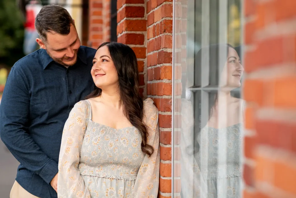 a couple stand and canoodle beside a brick wall and a window. Her reflection is visible in the window. Part of an engagement shoot Shot in Niagara-on-the-lake by Pixelesque Photography.