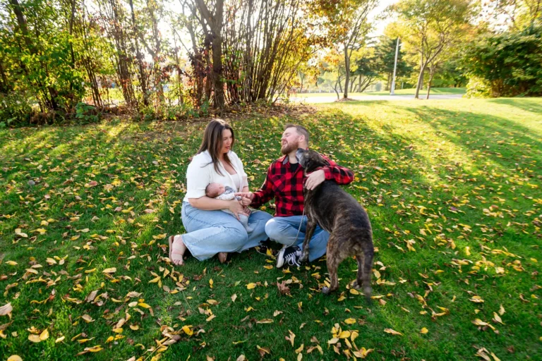 a dog licks a man as his wife and newborn sit beside him on the grass covered in fallen autumn leaves. Shot at bayfront park by Pixelesque photography - Hamilton Family Photographer