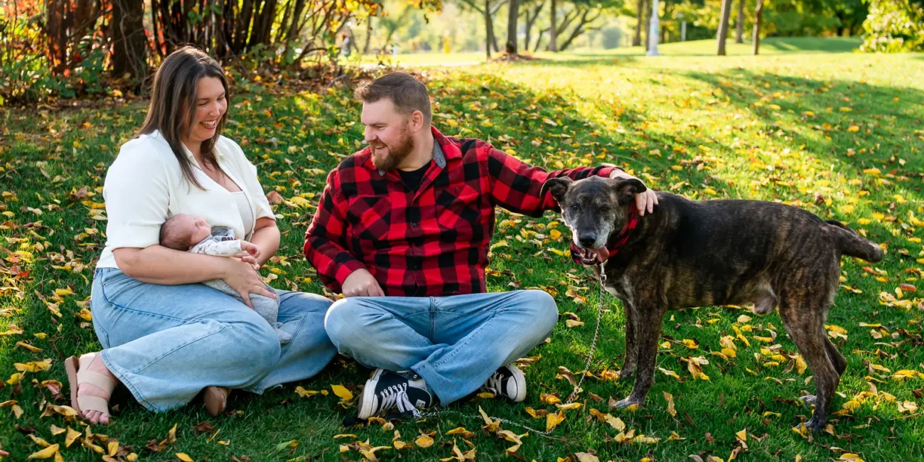 a mother holds her newborn in her lap. She, the father and their dog are sitting beside them on the grass covered in fallen autumn leaves. Shot at bayfront park by Pixelesque photography - Hamilton Family Photographer