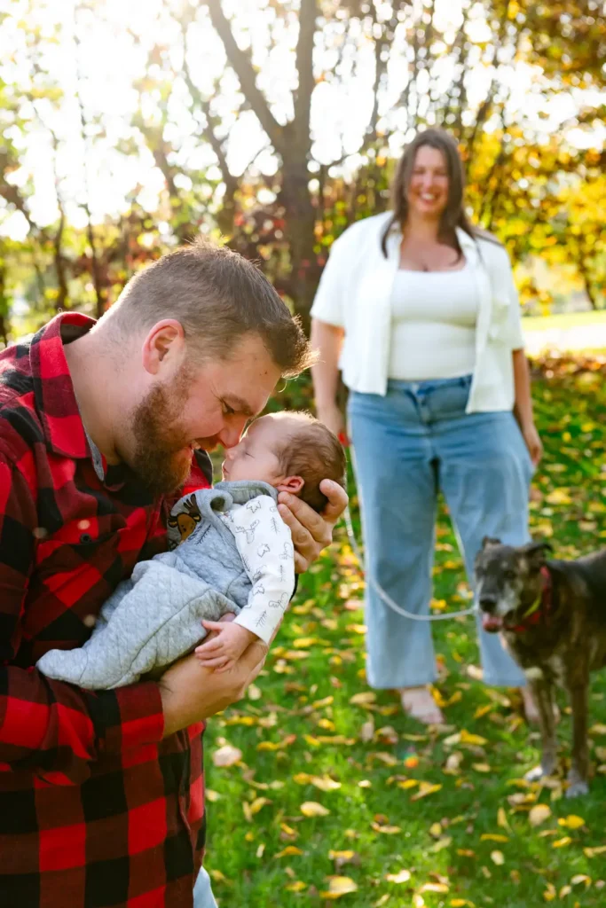 a man touches his nose to his newborn's nose as the mother and dog look on from behind. Shot at bayfront park by Pixelesque photography - Hamilton Family Photographer