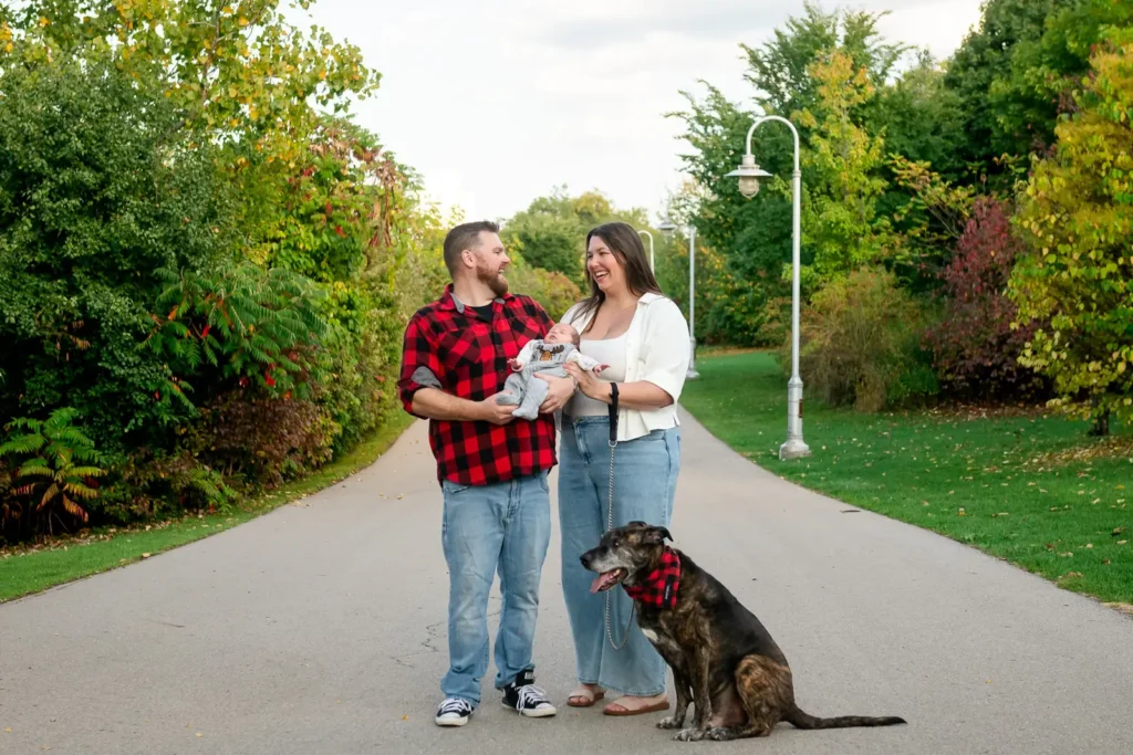 A couple hold their newborn baby and laugh towards each other and their dog sits by them. Shot at bayfront park by Pixelesque photography - Hamilton Family Photographer