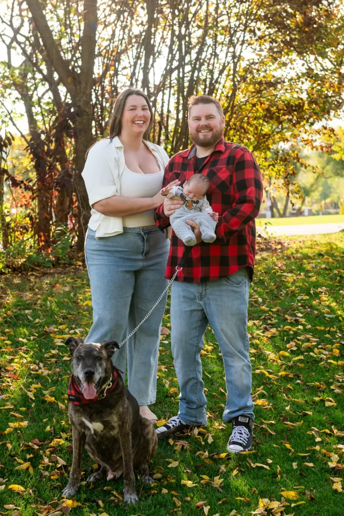 a couple hold their newborn baby and their dog sits on the ground at their feet. Shot at bayfront park by Pixelesque photography - Hamilton Family Photographer