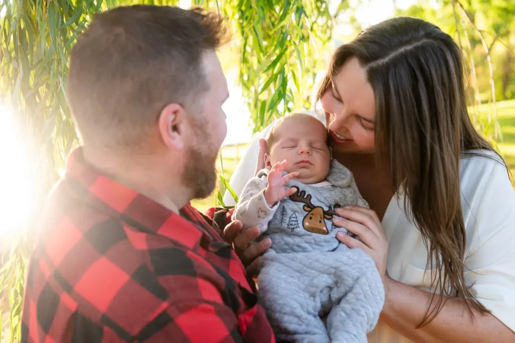 a couple holds their newborn up with sun bursting behind them and the willow tree. Shot at bayfront park by Pixelesque photography - Hamilton Family Photographer