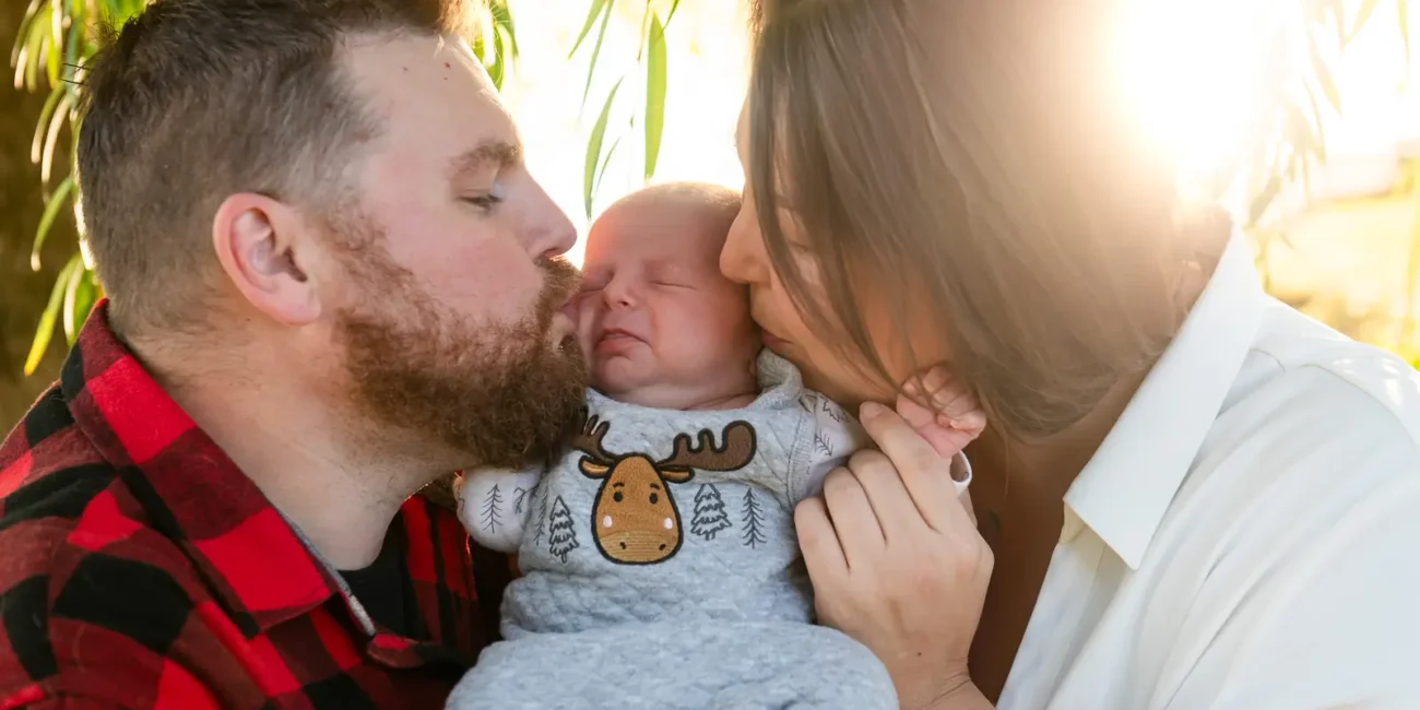a mother and father kiss their sleeping newborn on the cheeks as the sun bursts behind them. Shot at bayfront park by Pixelesque photography - Hamilton Family Photographer