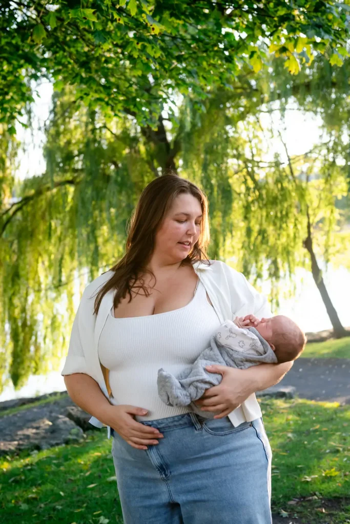 a mother holds her newborn on her hip with a large tree behind them. Shot at bayfront park by Pixelesque photography - Hamilton Family Photographer