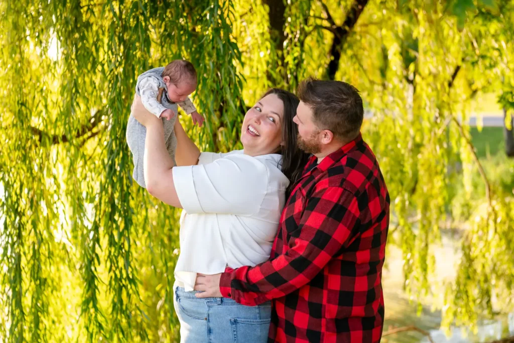 a mother holds up her newborn baby and looks back at her husband behind her as willow branches behind them are lit up by the sun. Shot at bayfront park by Pixelesque photography - Hamilton Family Photographer
