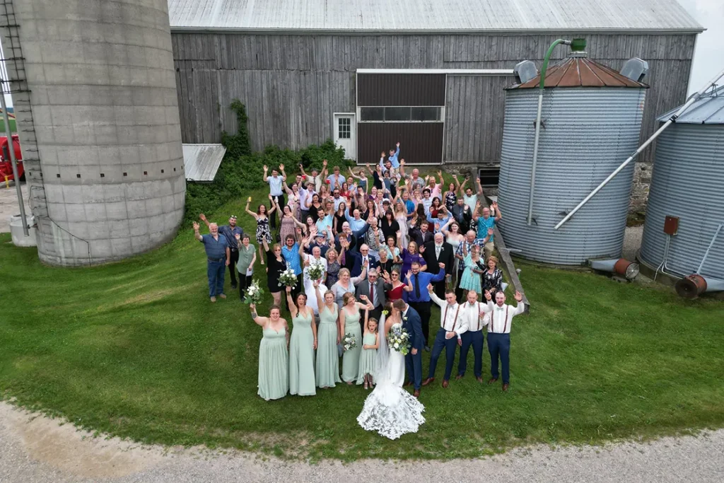 A photograph of the entire wedding guest list as the couple kiss in from of them and a barn. A wedding day drone photograph shot by Pixelesque Photography - Hamilton wedding Photographer.