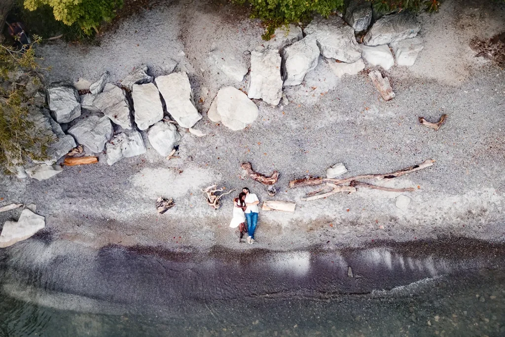 A couple lay together on a rocky beach as the waves gently crash by their feet at Brueckner Rhododendron Gardens. A wedding day drone photograph shot by Pixelesque Photography - Hamilton wedding Photographer.