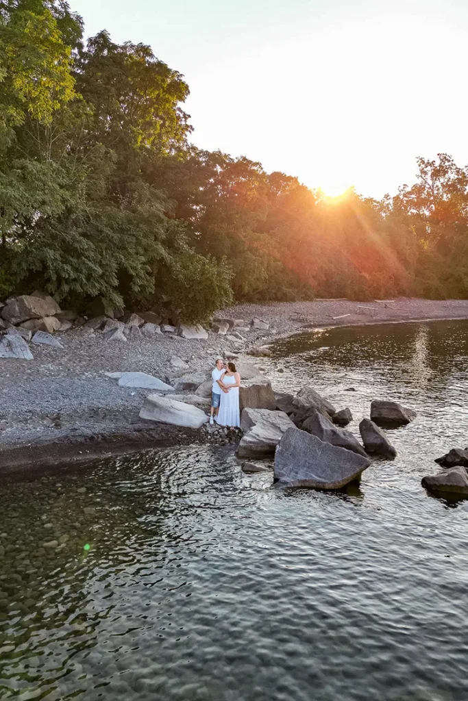 A couple share an embrace at shore of Lake Ontario as the sun sets behind them at confederations beach park. An engagement session drone photograph shot by Pixelesque Photography - Hamilton wedding Photographer.