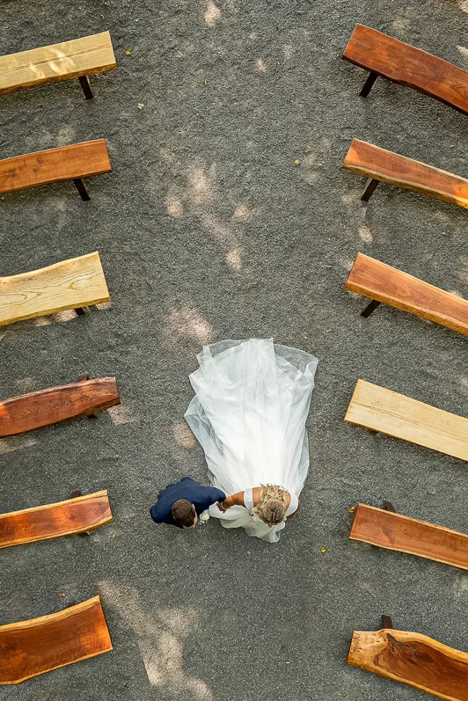 A couple walk down the aisle holding hands through empty wooden seats at CopeTown Woods golf club. A wedding day drone photograph shot by Pixelesque Photography - Hamilton wedding Photographer.